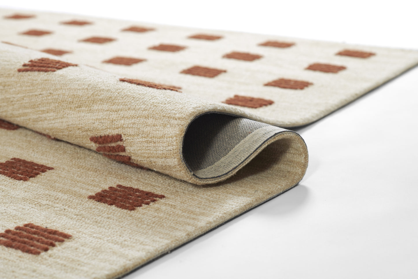Beige woven area rug with rust-brown square motifs, partially rolled to reveal gray underside.