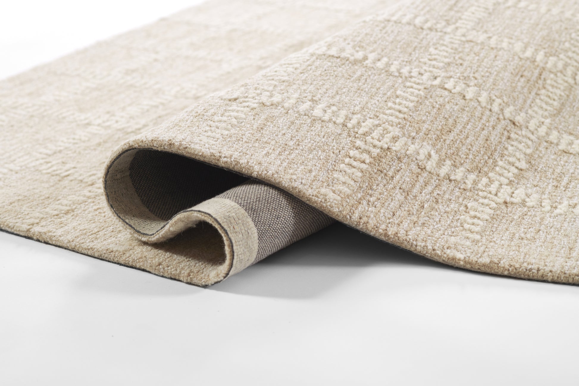 Beige patterned area rug with subtle geometric texture and medium plush pile, edge rolled to reveal gray backing.