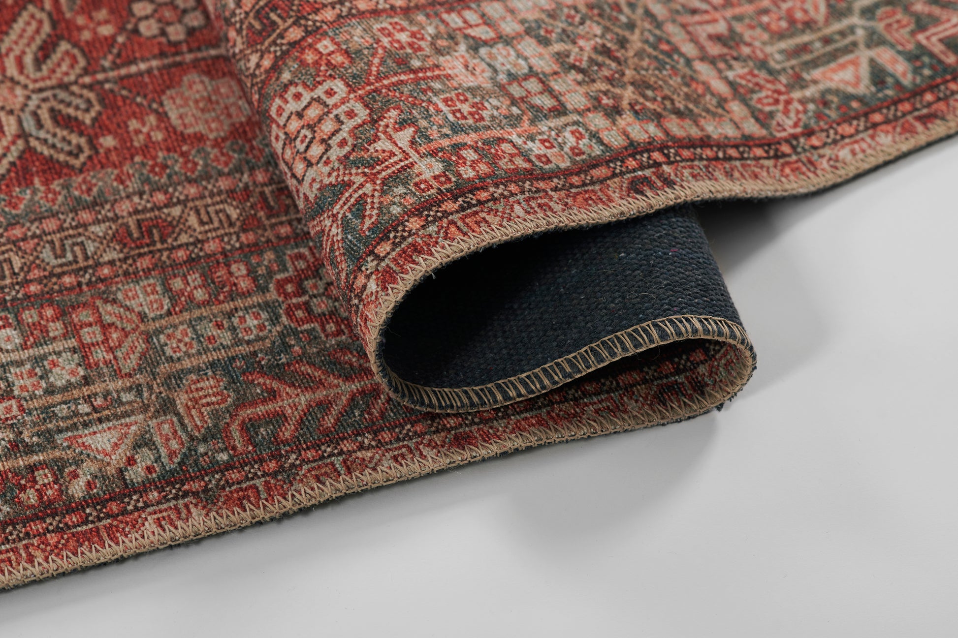 Red Persian-style rug with intricate geometric motifs, rolled edge showing beige stitching and dark backing.