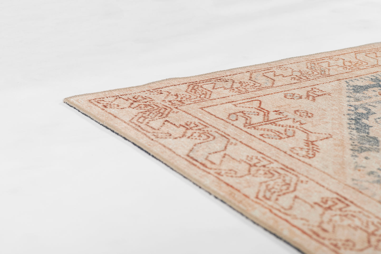 Corner view of a peach-beige patterned area rug with a geometric border and blue accents.