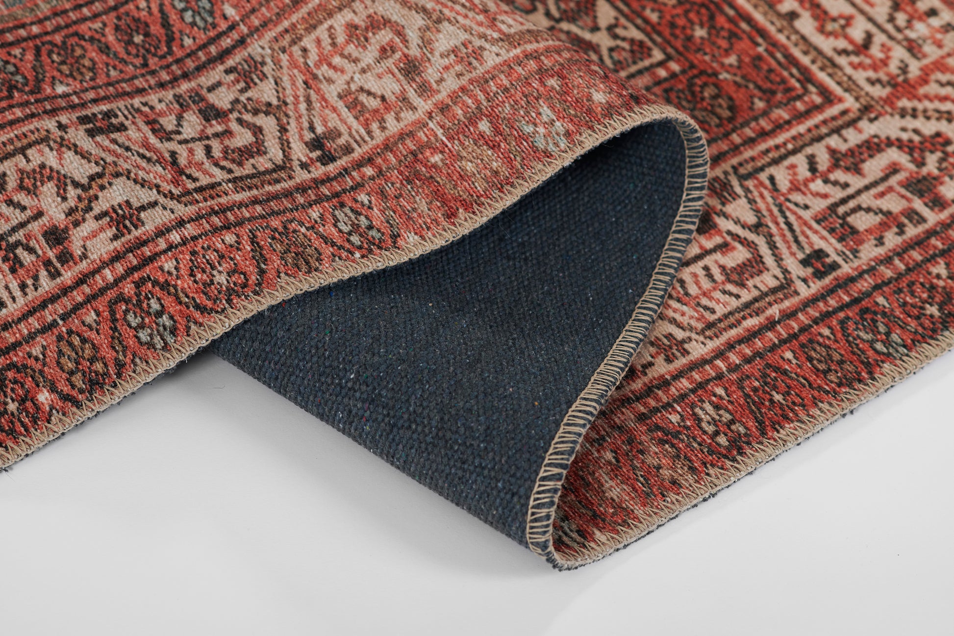 Traditional red Persian-style rug with ivory geometric motifs and fringed edges; rolled corner reveals navy blue underside.