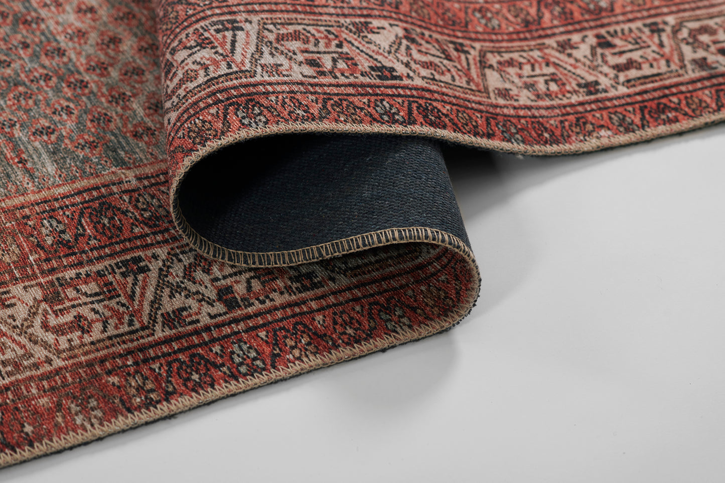 Red and beige Persian-style rug with intricate geometric borders; rolled corner reveals dark underside.