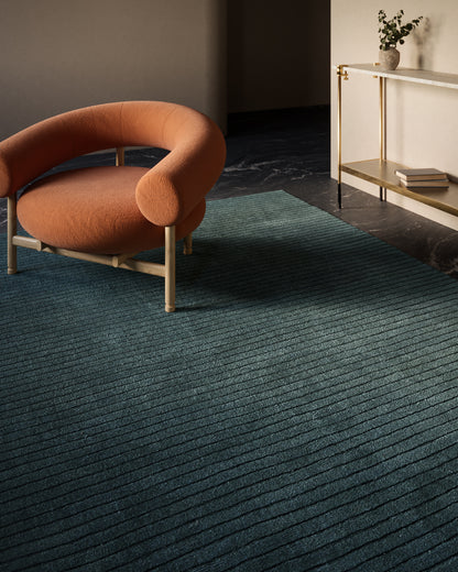 Orange boucle lounge chair with rounded back and light wood frame on teal striped rug.