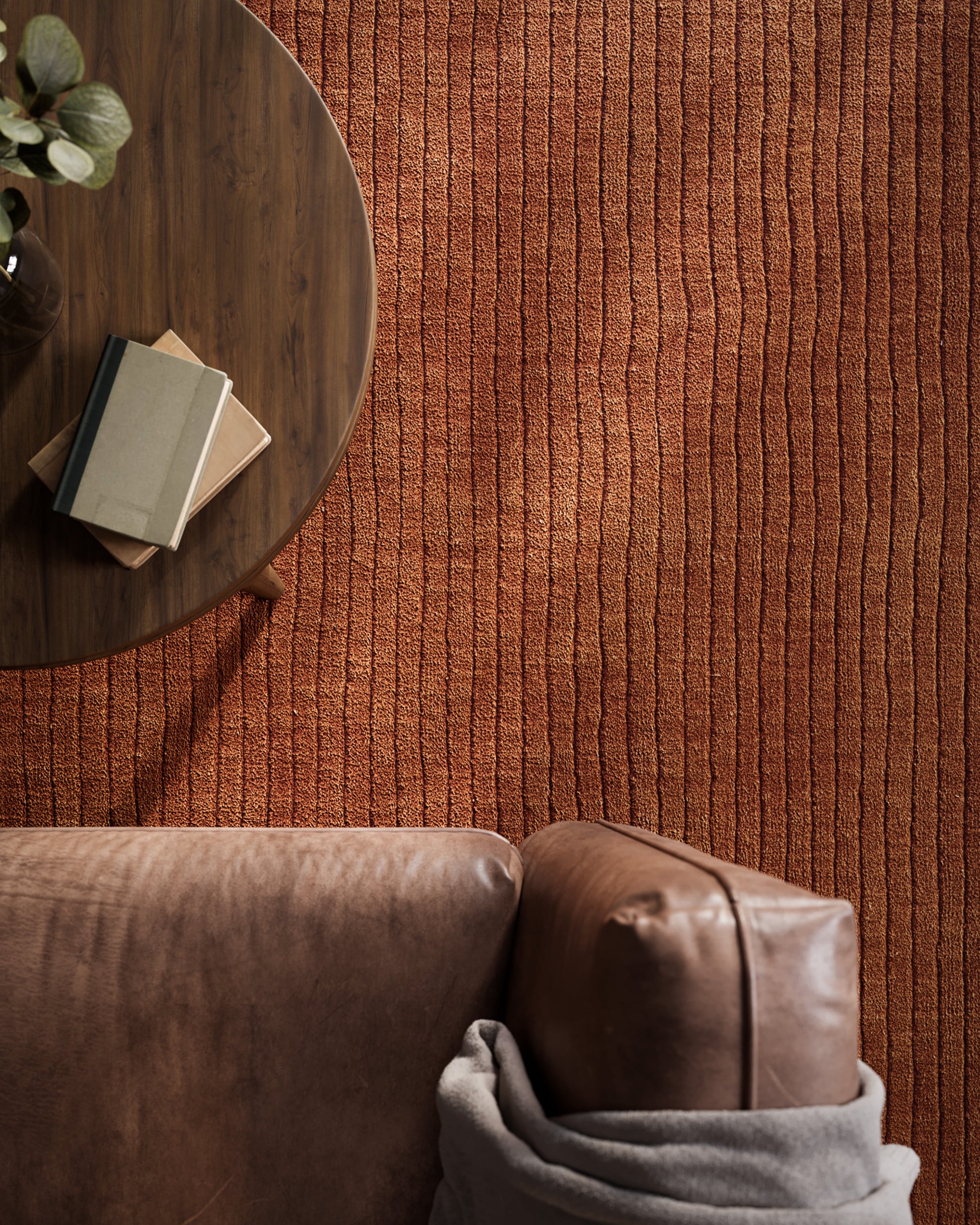 Cozy living room scene with round wooden coffee table, stack of books, potted plant, rust-orange rug, and brown leather sofa.