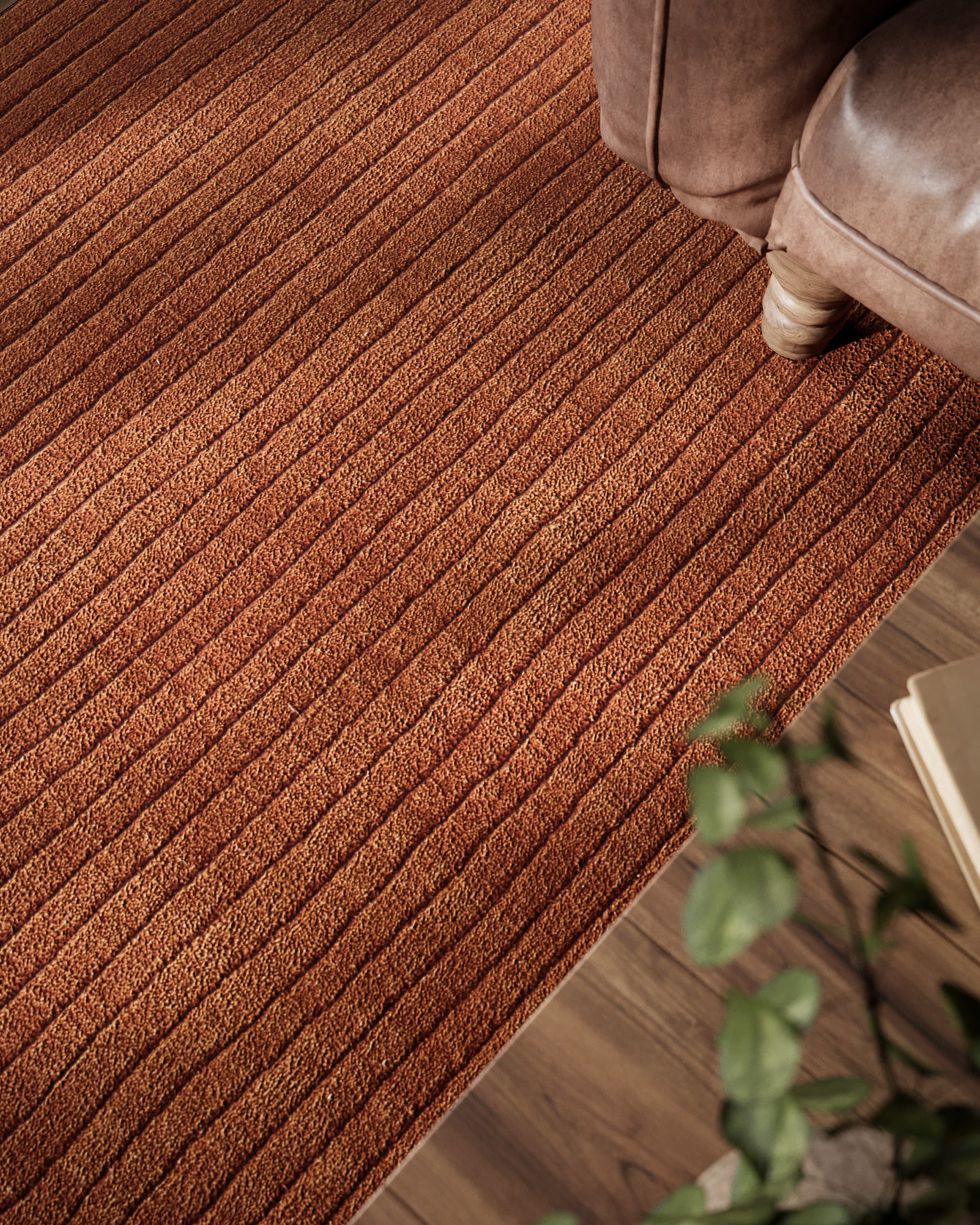 Rust-brown textured ribbed rectangular rug with striped loop pattern on wood floor beside a brown sofa and potted plant.