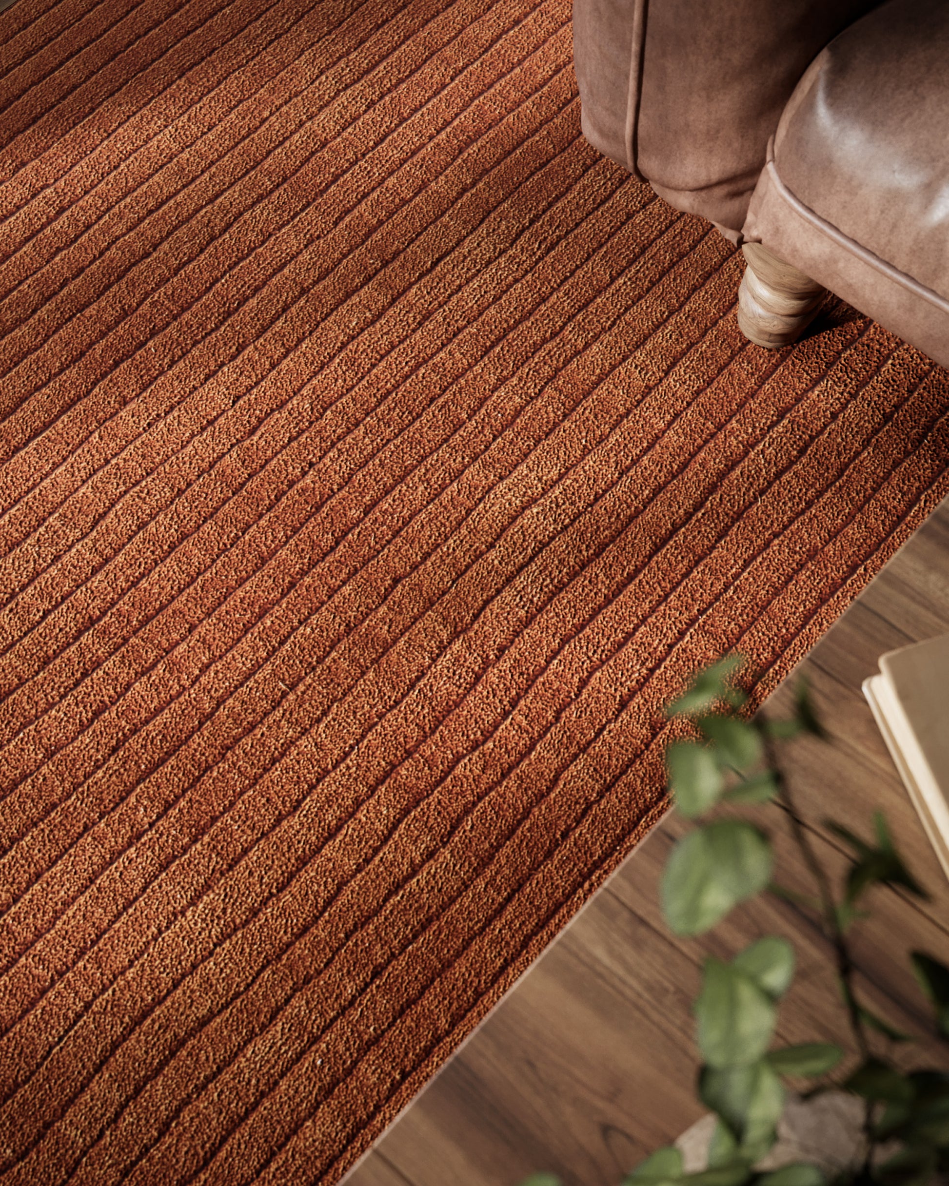 Rust-orange striped textured rug on wood floor beside a brown sofa, with a potted plant in the foreground.