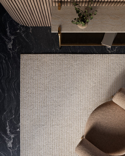 Top-down view of a beige ribbed rug on dark marble floor, tan sofa, and a marble-topped console with gold legs and a plant.