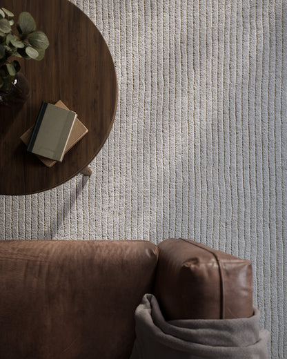 Round dark wood coffee table on pale striped rug beside a brown leather sofa with a gray throw; stacked books and a eucalyptus vase.