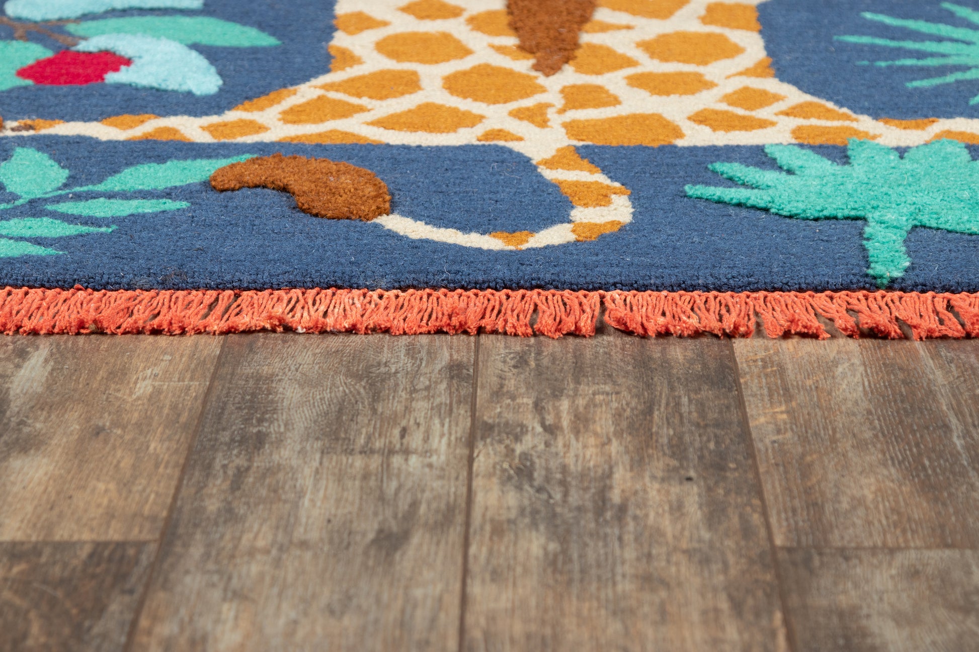 Blue tufted rug with orange giraffe and tropical leaf patterns, orange fringe along edge on a wooden floor.