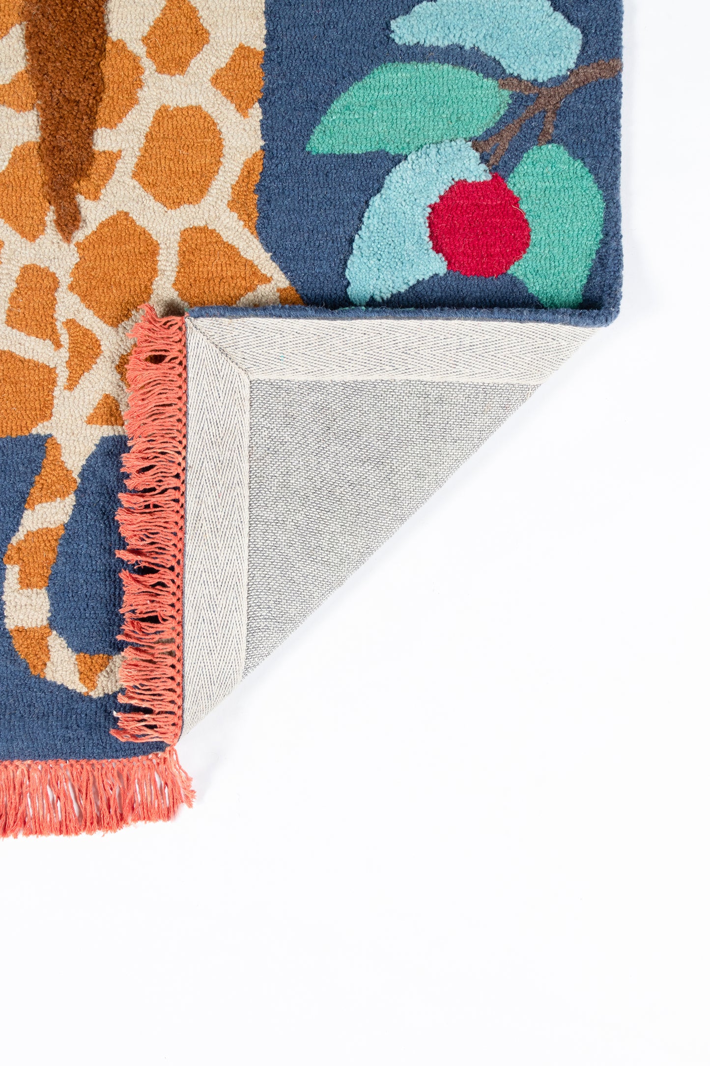 Colorful woven rug with navy blue and orange pattern, teal leaves, red berry, pink fringe; folded corner shows grey backing.