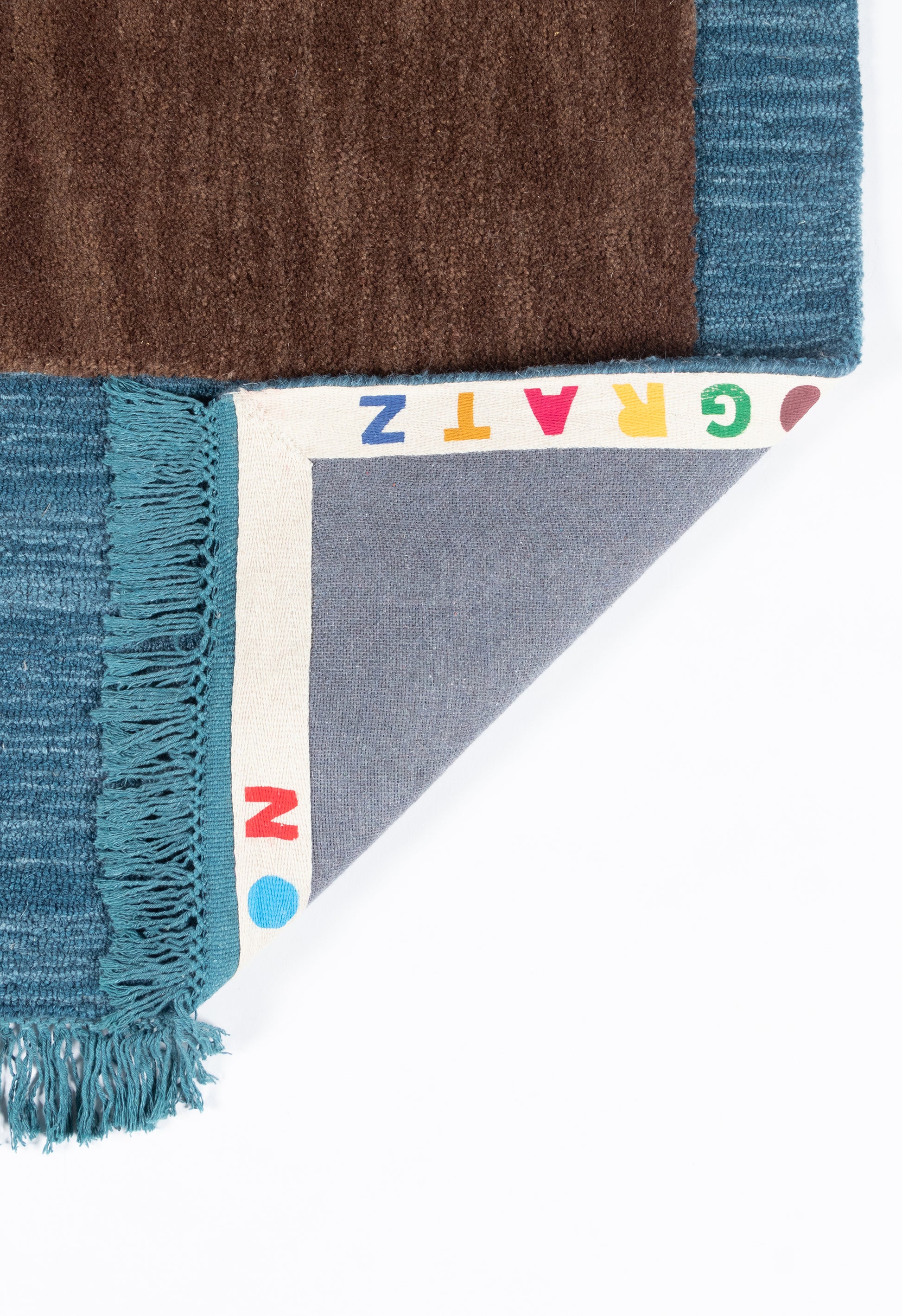 Turquoise-fringe rug sample with teal edge and brown swatch; folded corner shows gray backing and a rainbow-letter edge.