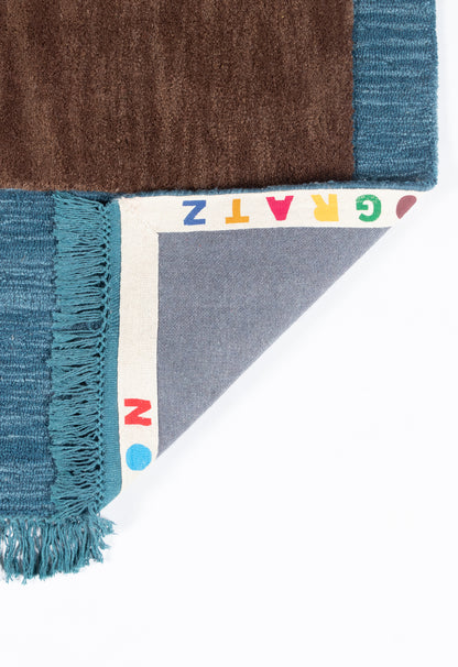 Turquoise-fringe rug sample with teal edge and brown swatch; folded corner shows gray backing and a rainbow-letter edge.