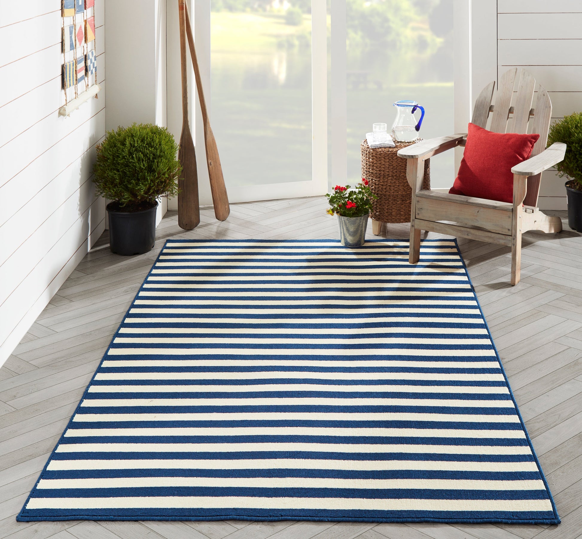 Blue and white striped outdoor rug on a light wood porch, with potted plants and a wooden Adirondack chair with a red cushion.