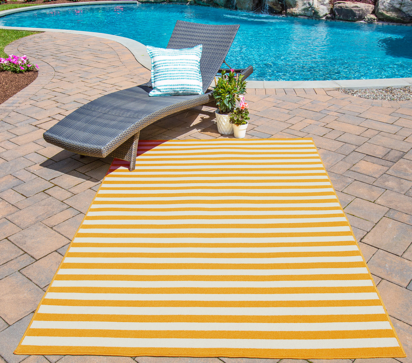 Yellow and white striped outdoor rug on brick patio beside a gray wicker lounge chair with blue-striped pillow near pool.