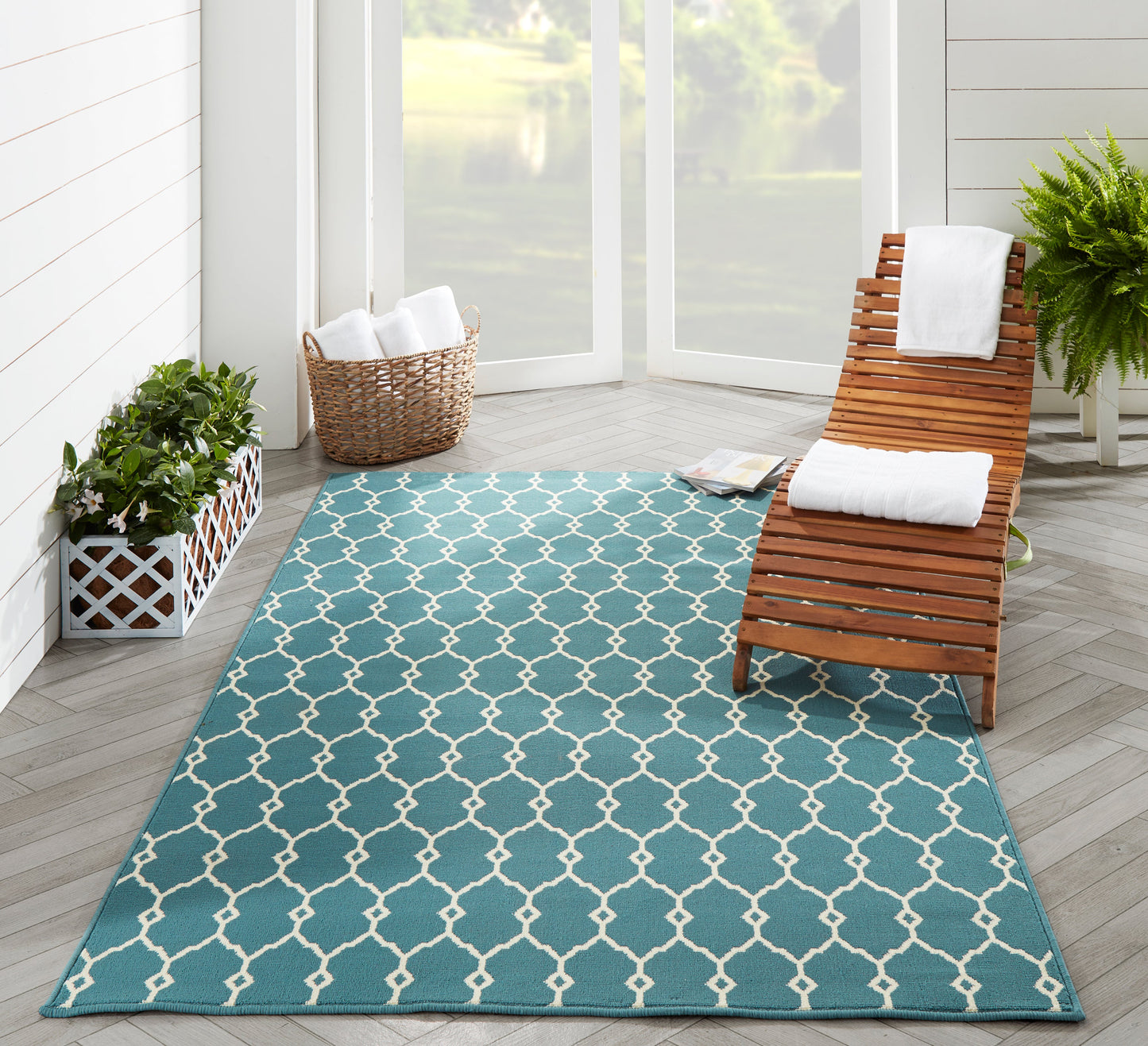 Teal outdoor rug with white lattice pattern on light wood deck, beside a wooden lounge chair and potted plants.