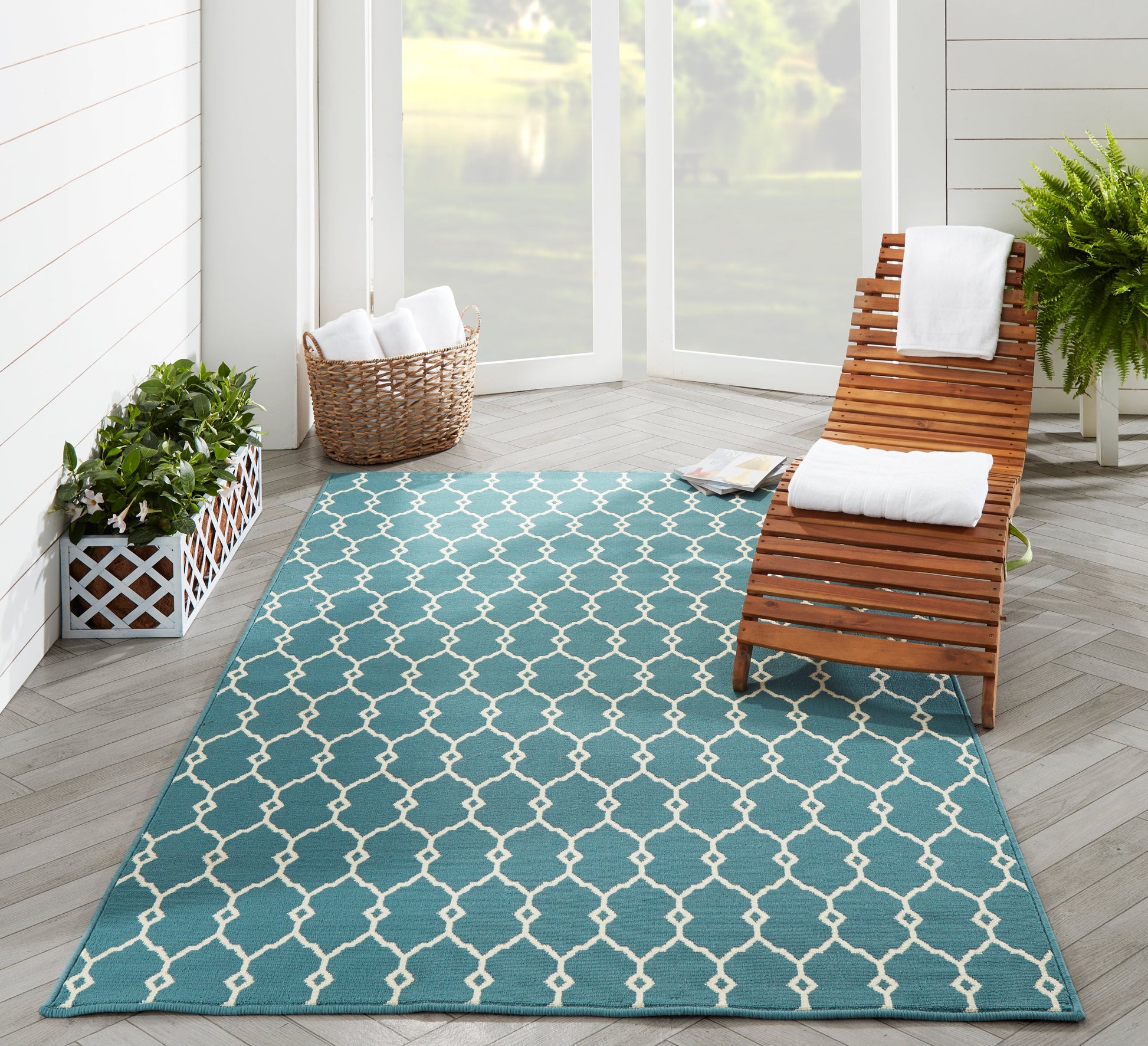 Teal outdoor rug with white lattice pattern on light wood deck, beside a wooden lounge chair and potted plants.