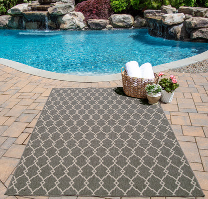 Gray outdoor rug with geometric lattice pattern on poolside brick patio, basket of white towels and potted plants nearby.