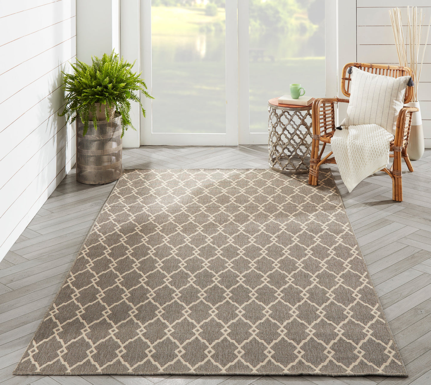 Taupe area rug with a white lattice trellis pattern in a bright sunroom, paired with a wicker chair and potted plant.