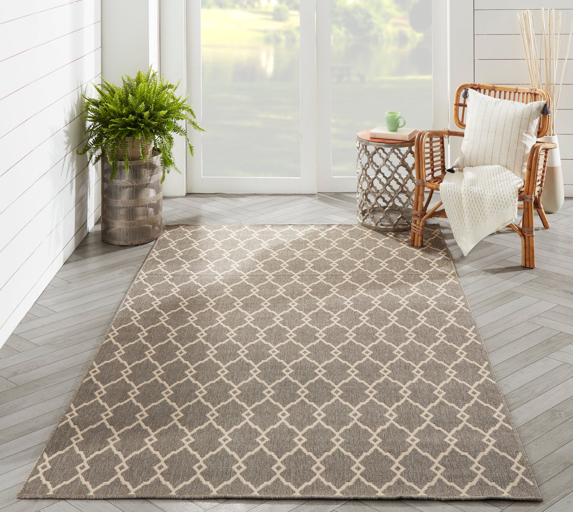 Taupe area rug with a white lattice trellis pattern in a bright sunroom, paired with a wicker chair and potted plant.