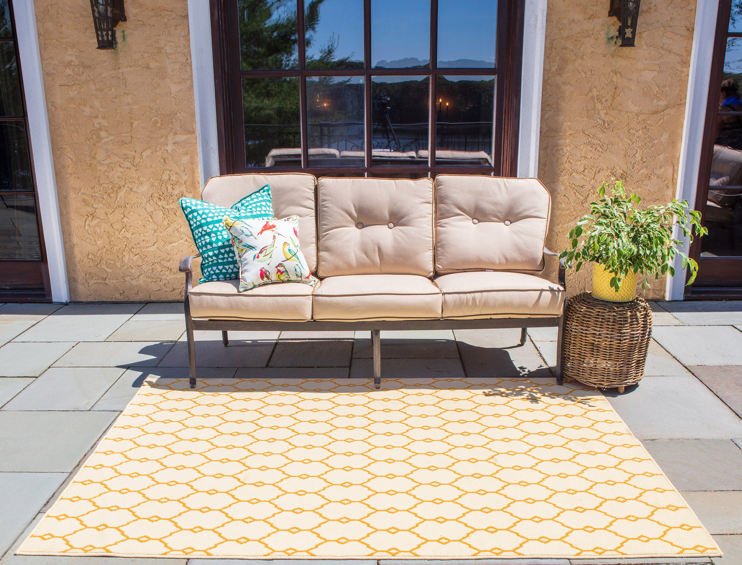 Beige outdoor sofa with metal frame, teal floral cushions, on a yellow honeycomb rug beside a wicker plant basket.