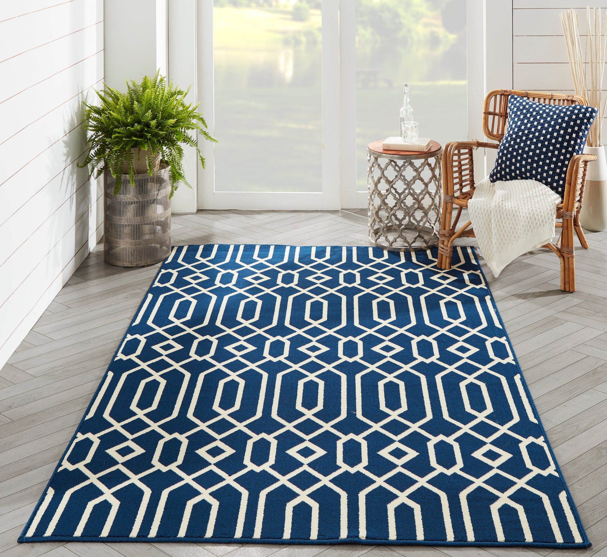 Navy blue area rug with white geometric lattice pattern in a sunlit living room; potted plant and wicker chair nearby.