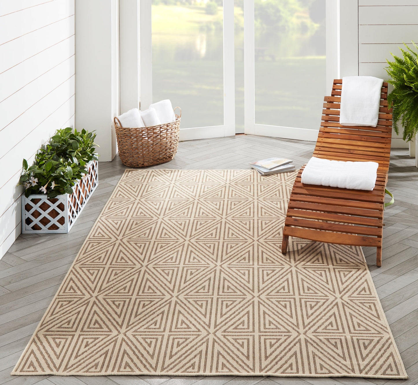 Geometric taupe rug on grey wood floor, with a wooden slatted lounge chair, white towels, wicker basket, and greenery.