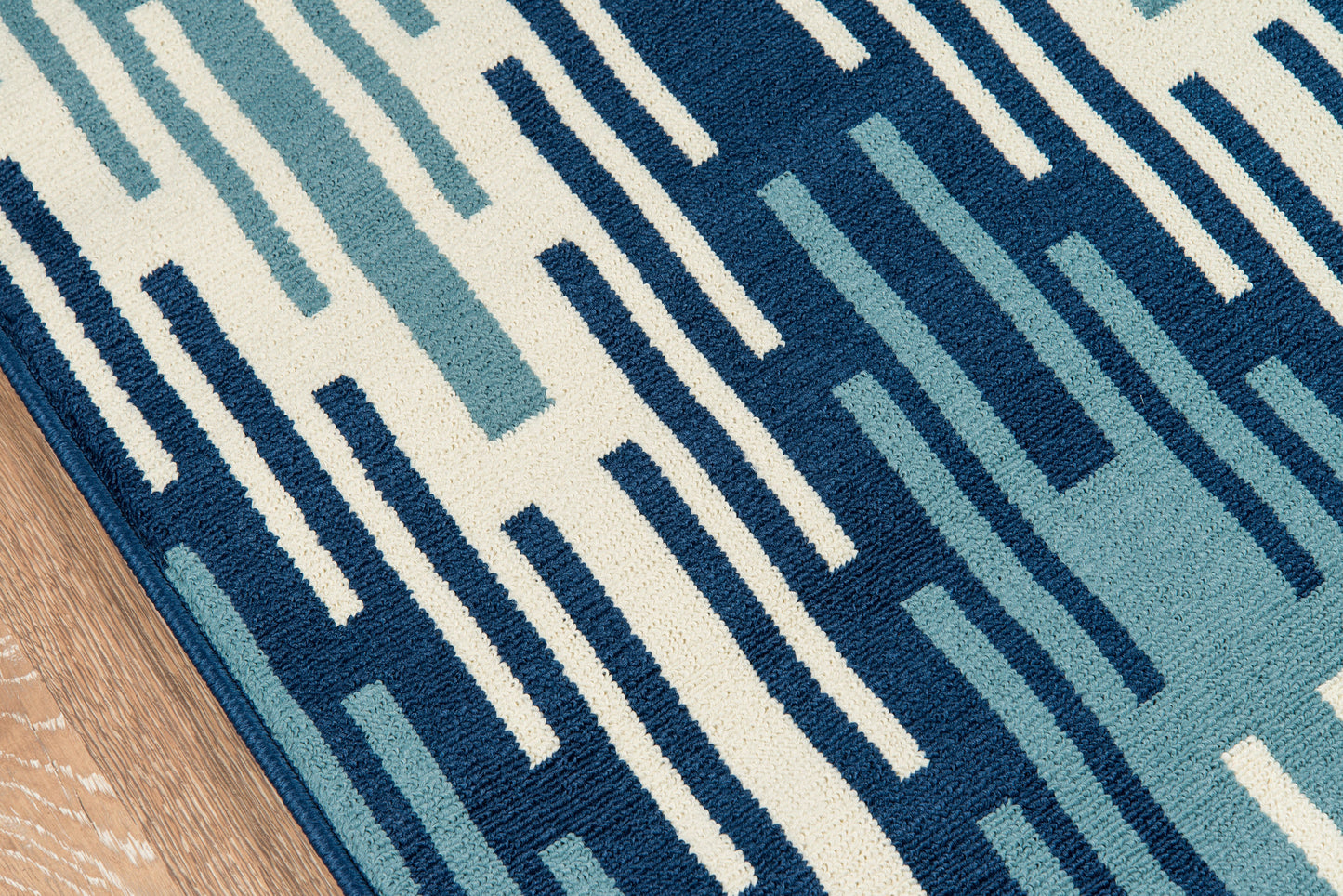 Geometric area rug with navy, blue and teal rectangular stripes on a cream background, edge visible on a wooden floor.