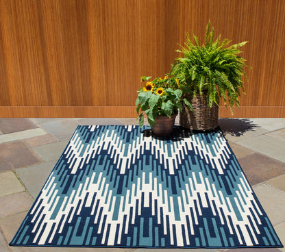 Rectangular outdoor rug in navy and white with a bold zigzag pattern on a stone patio beside sunflowers and a fern.