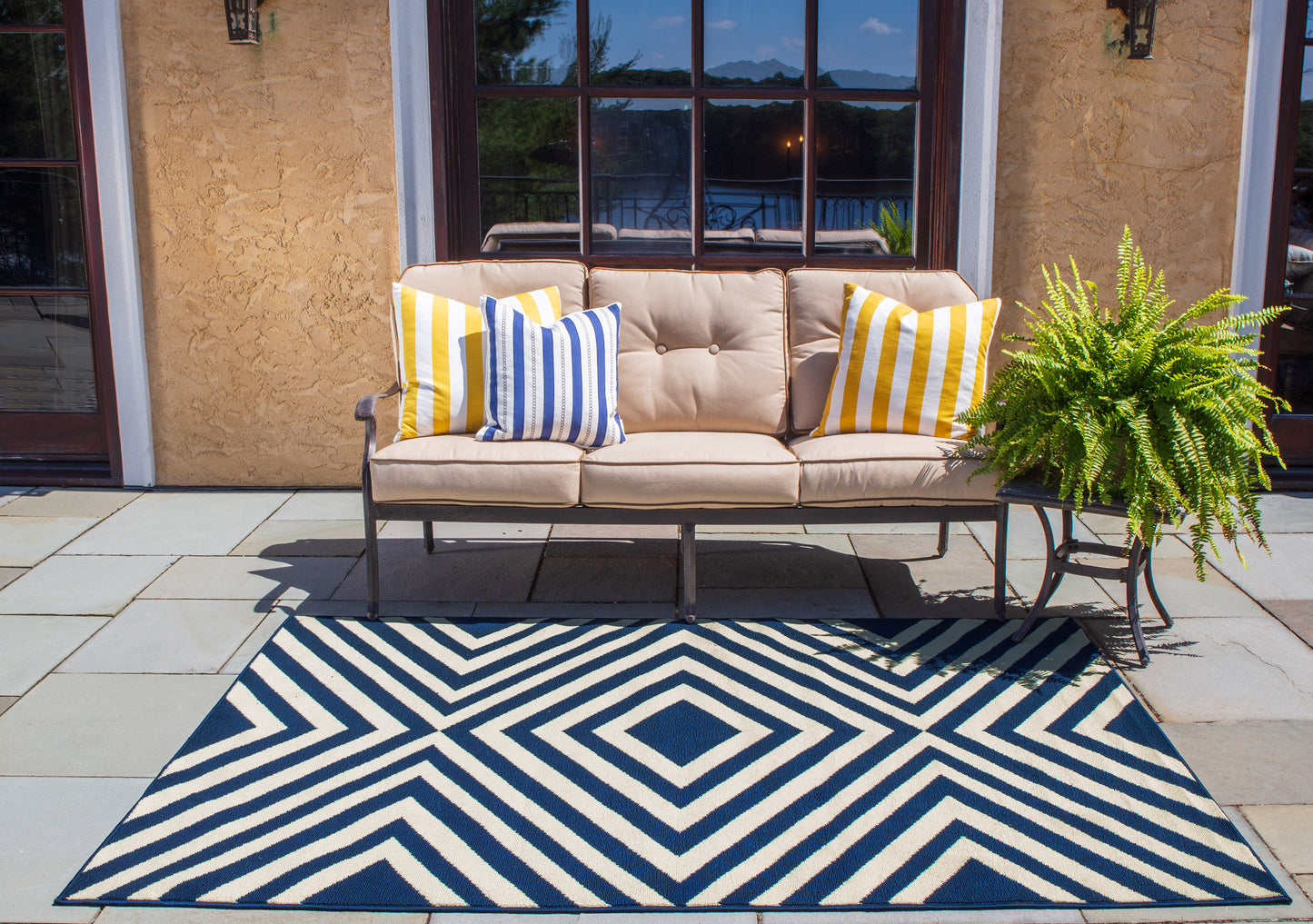 Navy and white geometric rug on a patio scene with a beige patio sofa with a metal frame, yellow and blue striped cushions, and a fern on the side table.