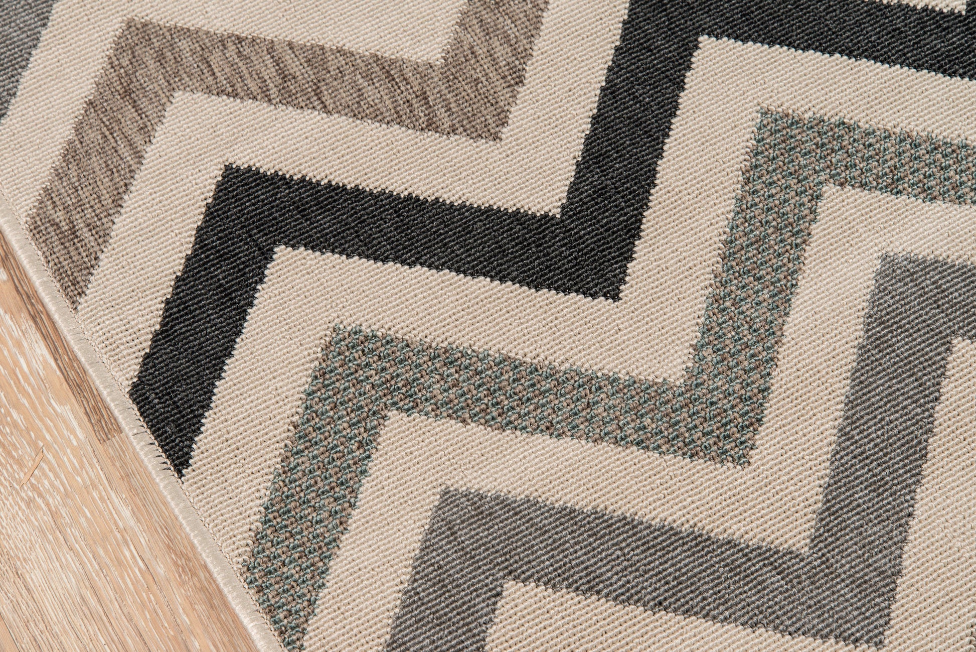 Beige woven area rug with a bold geometric zigzag pattern in black, brown, gray and teal on a wood floor.