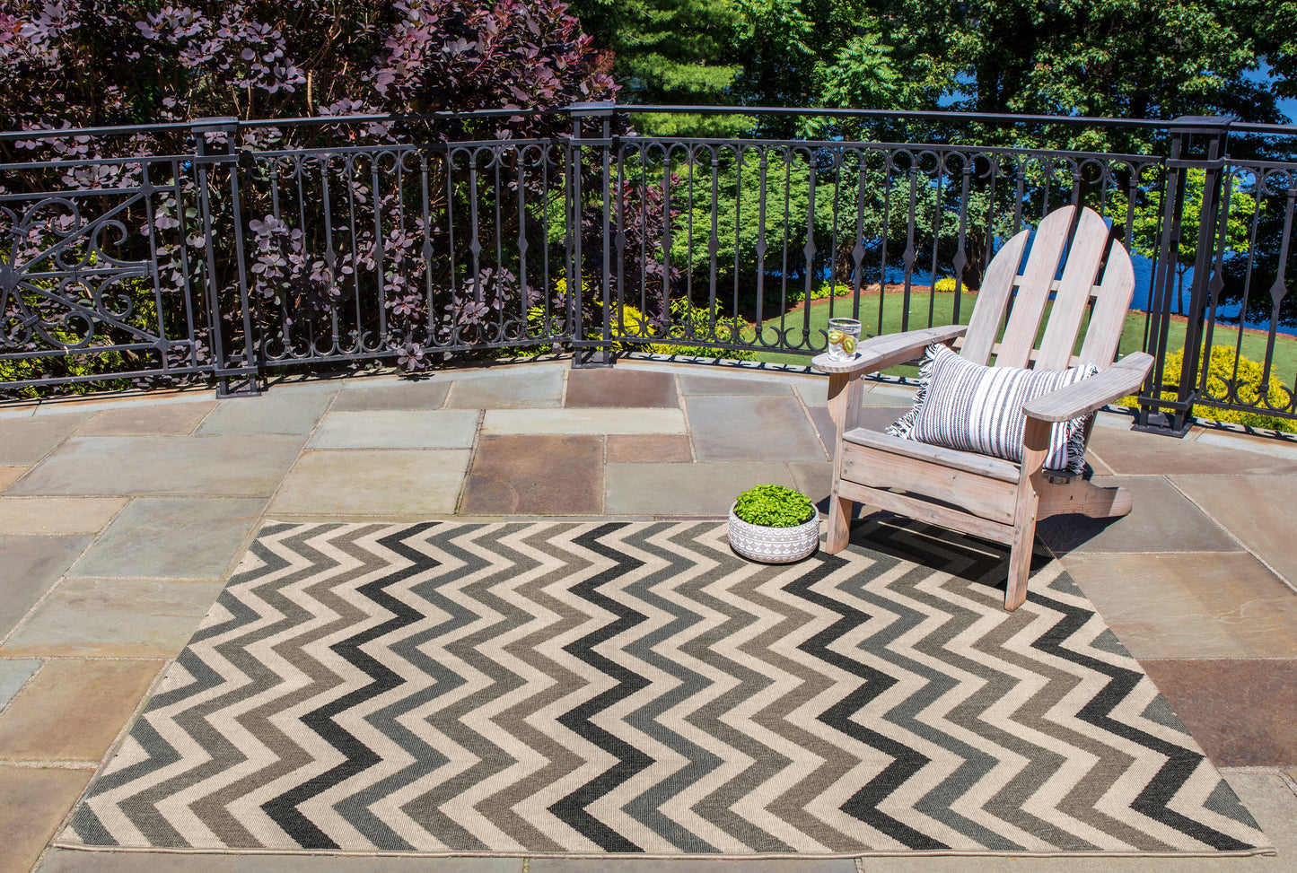 Outdoor patio with tan and gray chevron rug, weathered wood Adirondack chair, striped cushion, metal railing, potted plant.