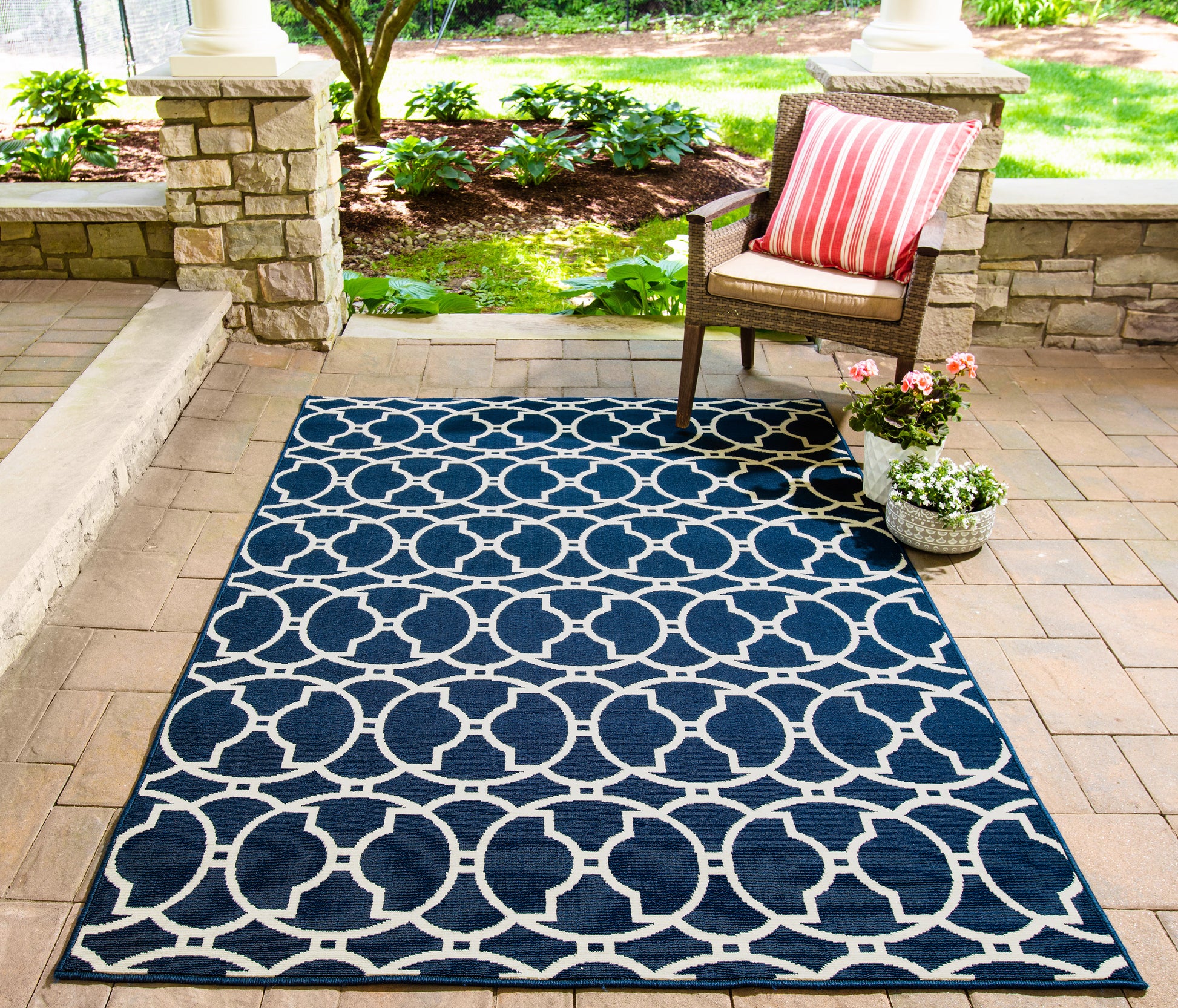 Navy outdoor rug with white circular lattice pattern on a stone-patio, with a wicker chair, striped cushion, and potted flowers.