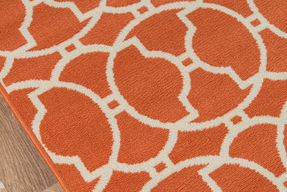 Orange rug with white interlocking circular lattice pattern and tufted texture on a wooden floor edge.