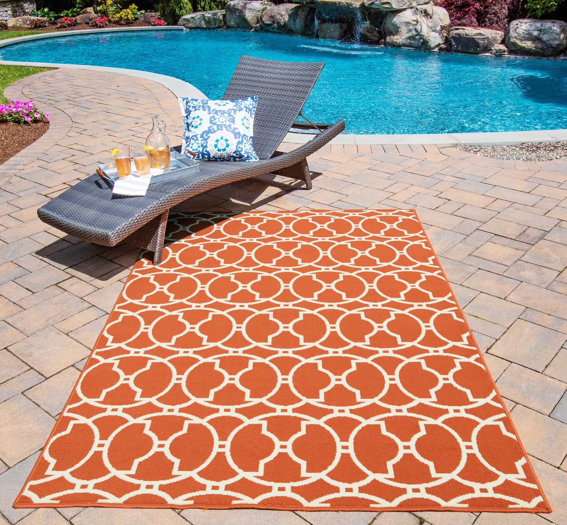 Outdoor poolside orange geometric area rug on brick patio beside brown wicker chaise lounge with blue patterned cushion.