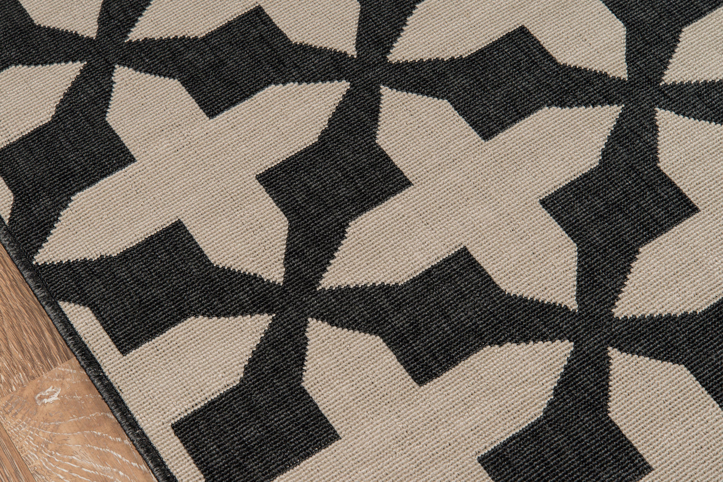 Beige and black woven rug with bold star-like geometric pattern; textured flatweave on wood floor edge.