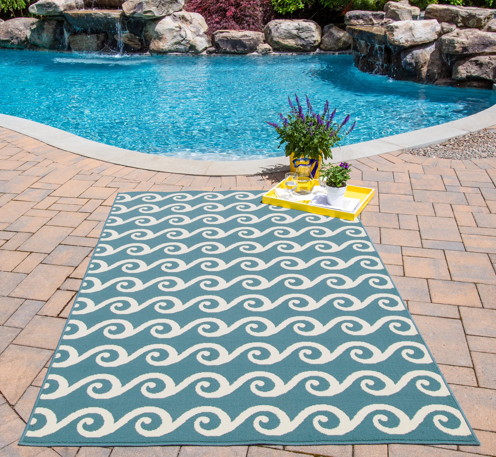 Rectangular outdoor rug in teal with white wave motif on brick poolside patio; yellow tray with potted lavender.