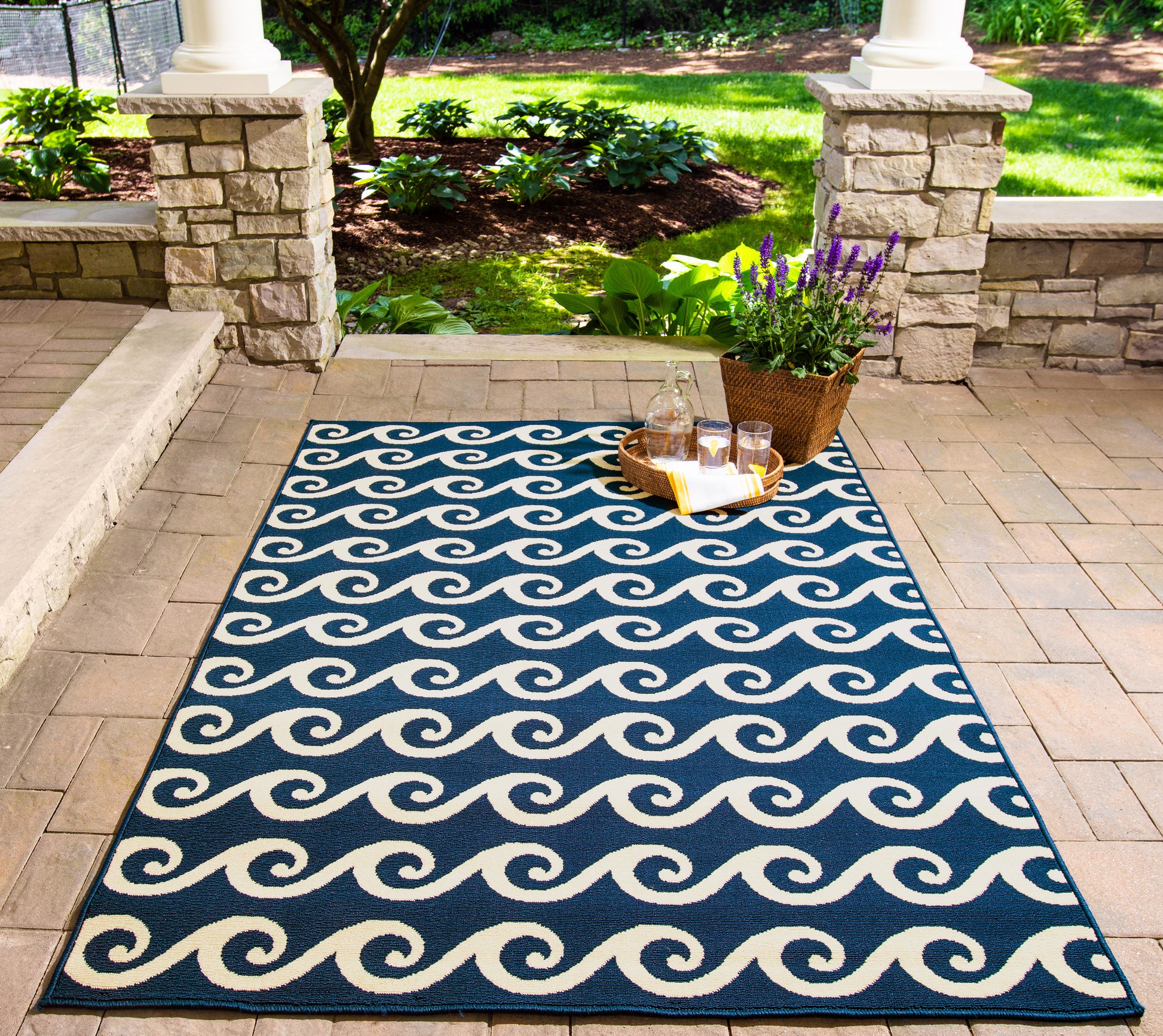 Navy blue outdoor rug with white wave pattern, rectangular on brick patio; wicker basket with lavender and drinks nearby.