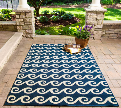 Navy blue outdoor rug with white wave pattern, rectangular on brick patio; wicker basket with lavender and drinks nearby.