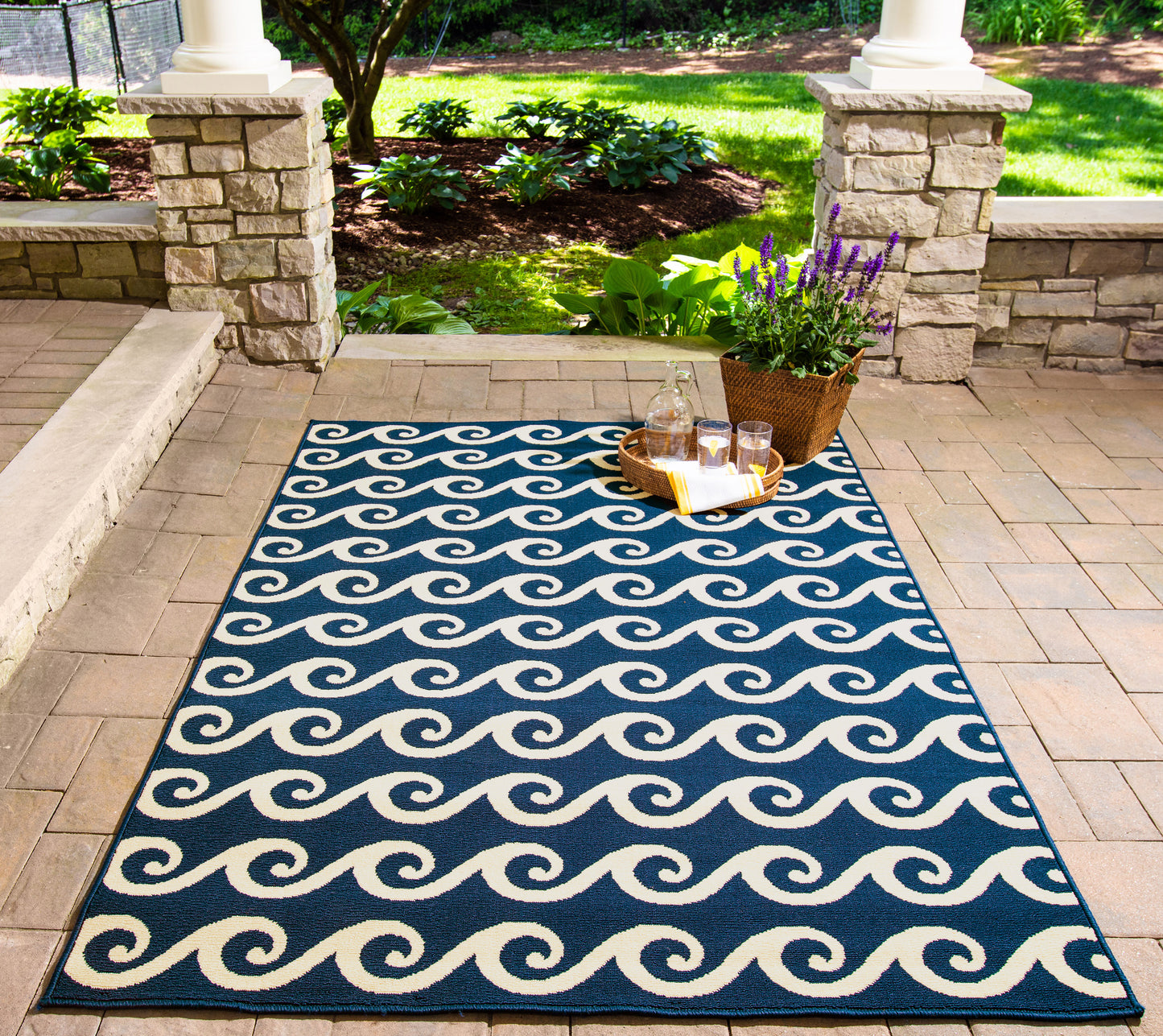 Blue outdoor rug with white wave pattern on brick patio; tray of drinks and a lavender plant in a wicker basket nearby.