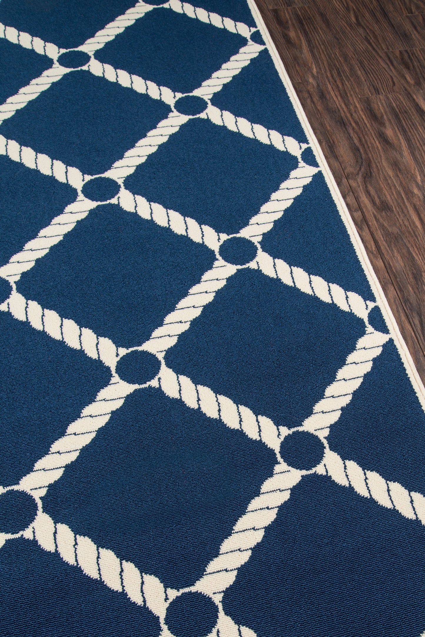 Blue area rug with white rope lattice forming diamonds and circular nodes; coastal geometric rug on dark wood floor.