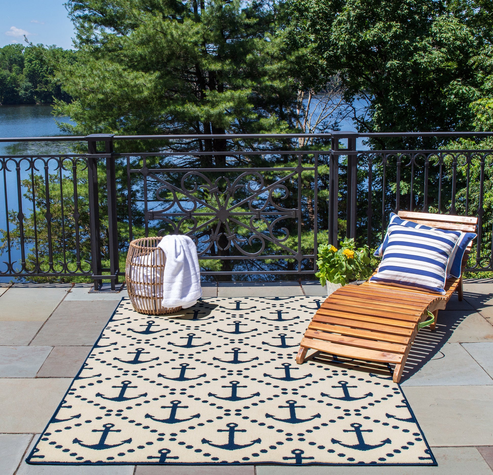 Nautical outdoor lounge: beige anchor rug, wooden chaise, navy-white striped cushion, wicker table, towel, yellow flowers.