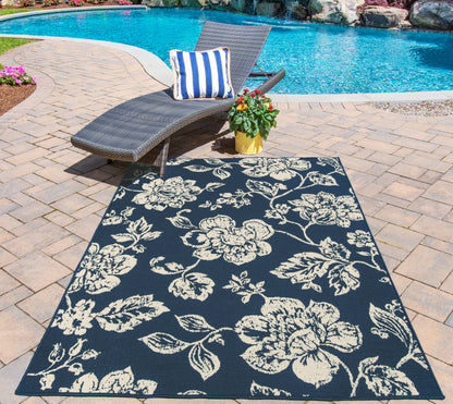 Poolside scene with navy floral outdoor rug, wicker lounge chair, blue & white striped pillow, potted plant on brick patio.