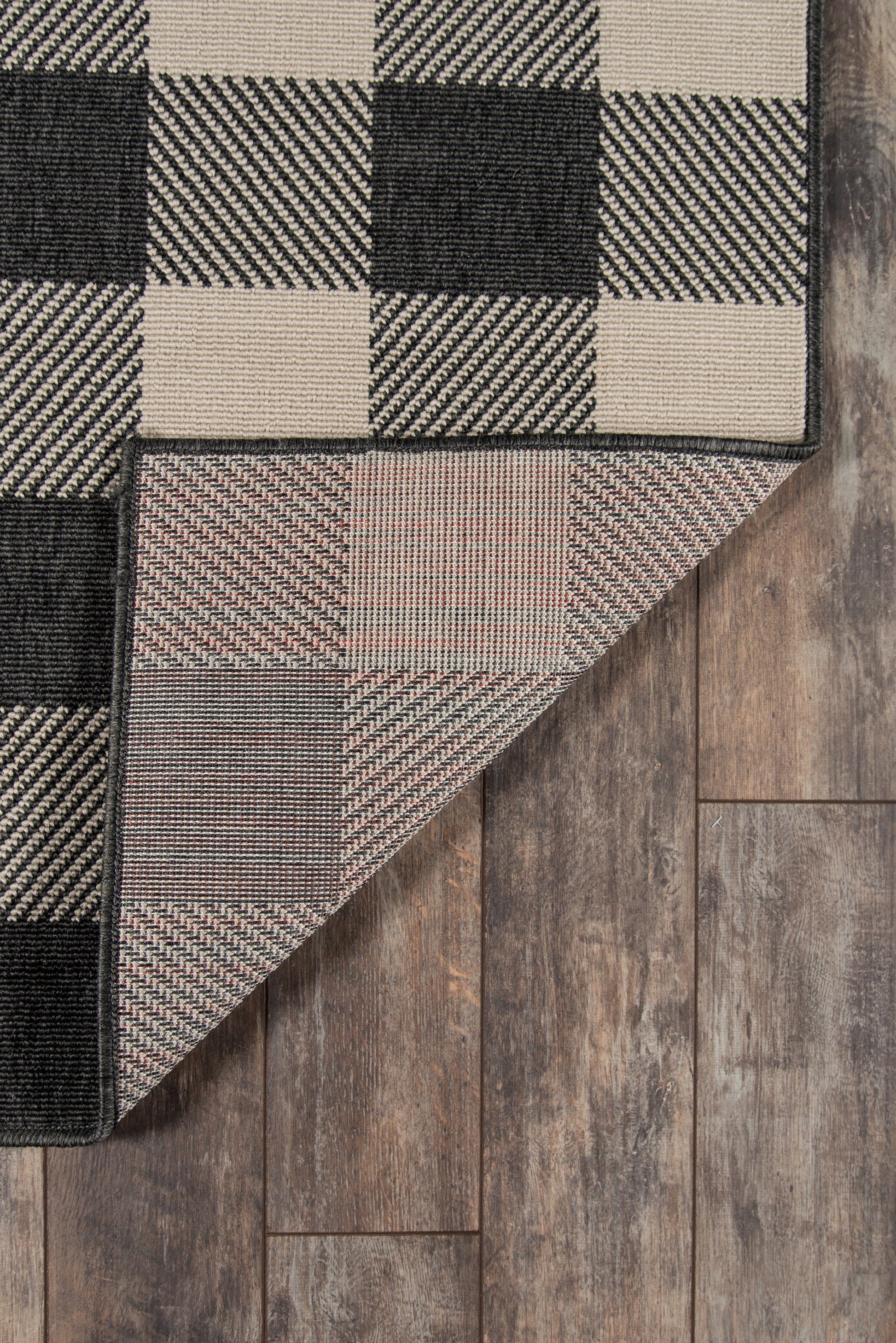 Woven checkerboard rug in black, beige and tan, folded corner reveals underside on wood floor.