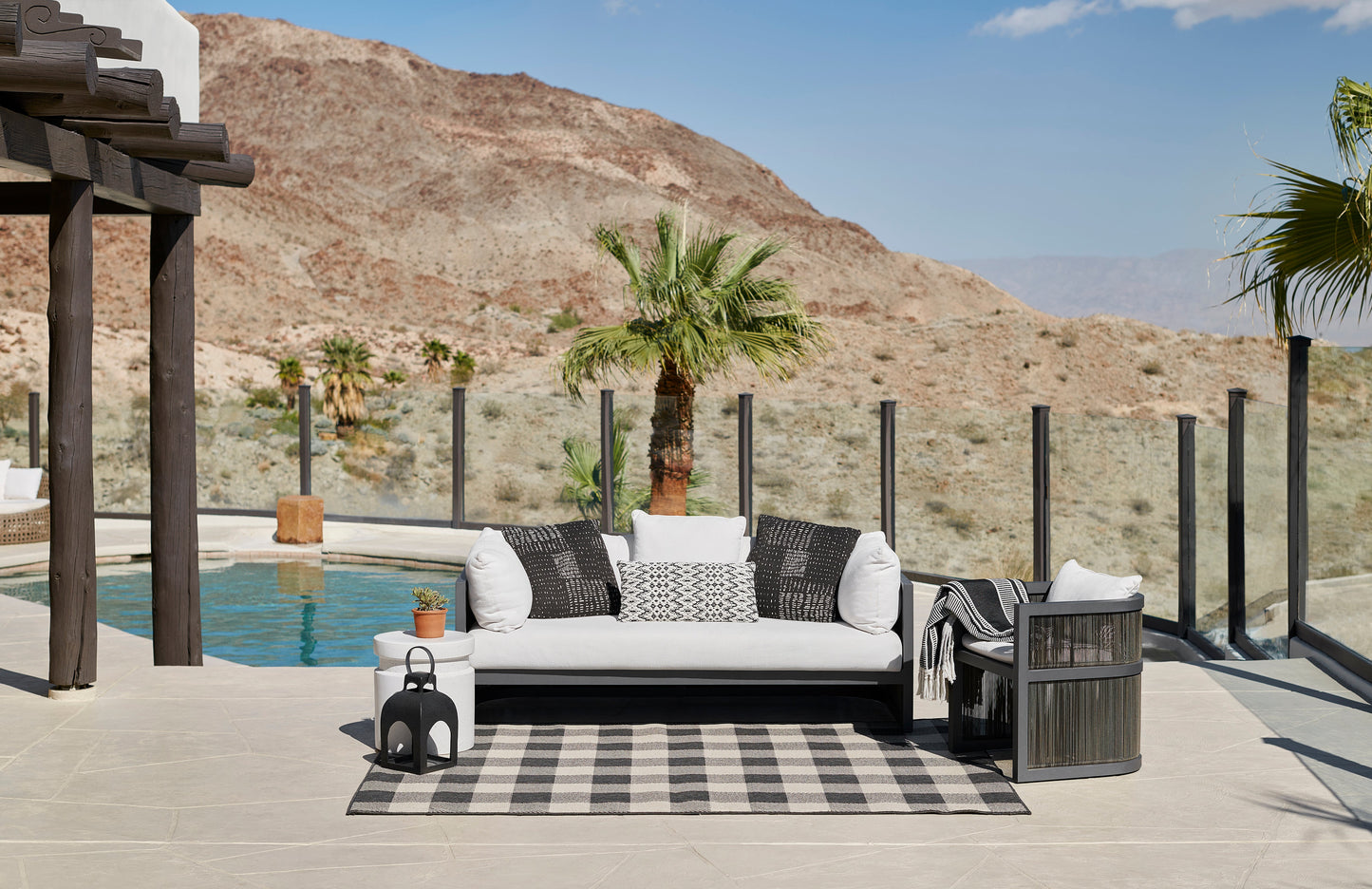 Modern outdoor lounge with white cushioned sectional, striped rug, palm trees, lanterns, glass railing, desert hills & pool.