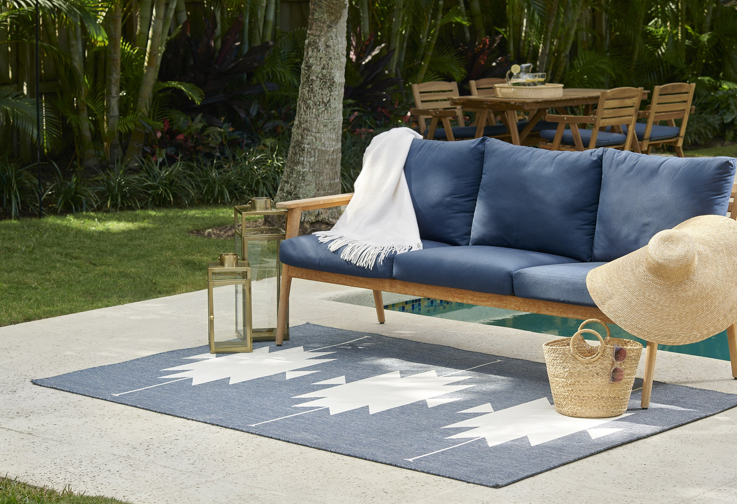 Outdoor wooden sofa with blue cushions, gray zigzag rug, straw hat, basket, lantern, and white throw blanket in garden patio.