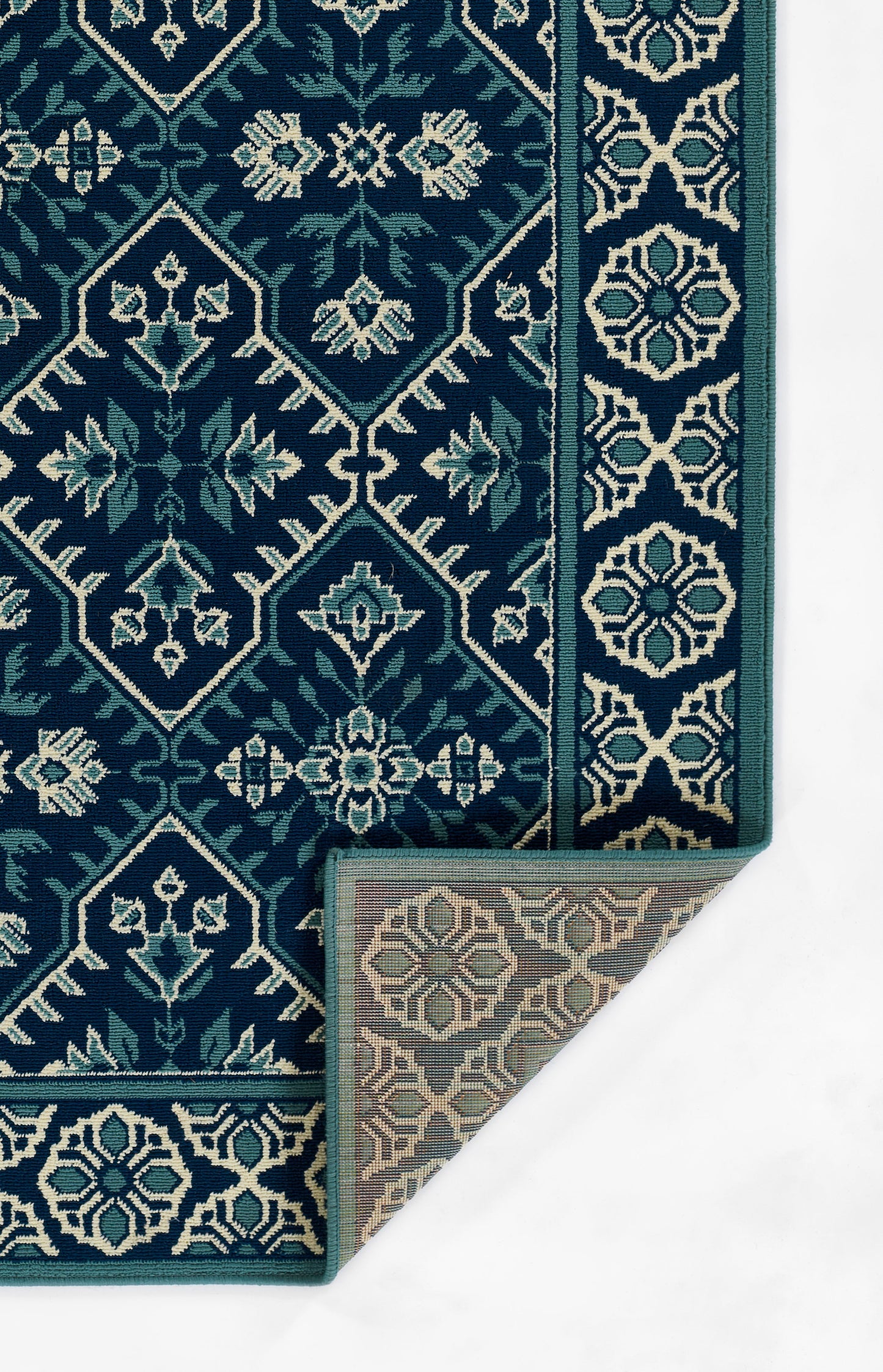 Dark teal woven rug with ivory geometric floral motifs and a bordered edge; folded corner reveals a beige underside.