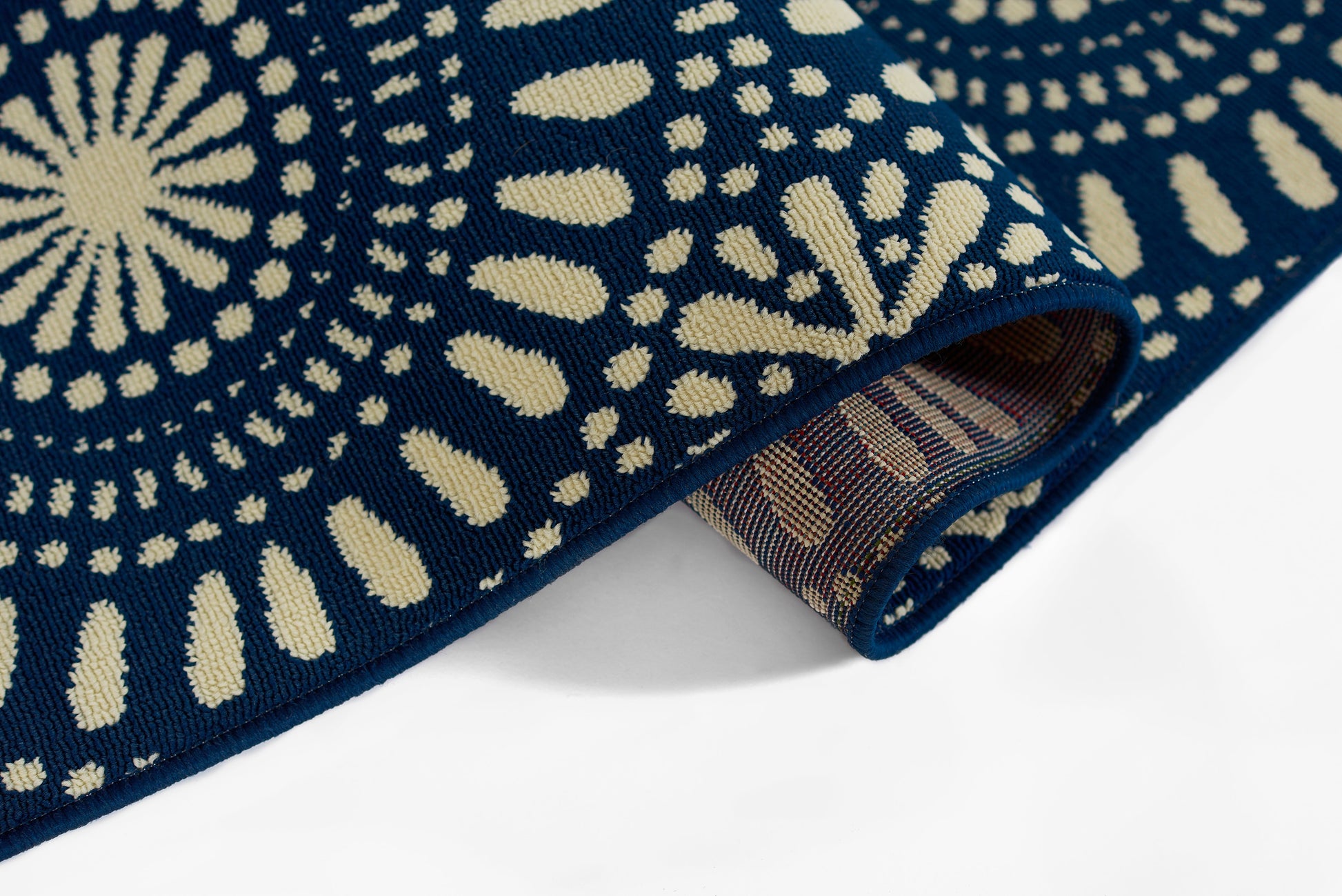 Navy blue woven rug with cream sunburst and dot motifs, rolled corner reveals brown striped underside.