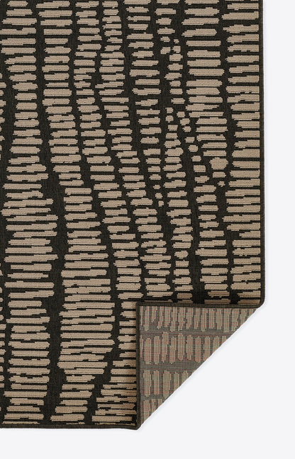 Rectangular rug with black background and irregular beige horizontal dashes; textured weave, folded corner reveals underside.