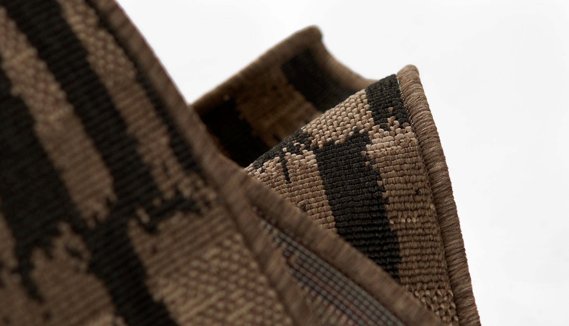 Close-up of brown and black woven rug with a geometric camo-like pattern; folded rug edge visible.