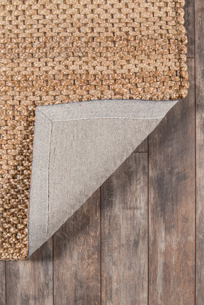 Natural braided jute rug with chunky weave; folded corner reveals gray fabric backing on wood floor.